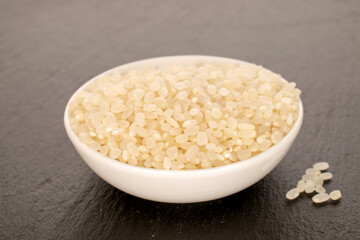 Round grain uncooked rice with white ceramic saucer on slate stone, macro.