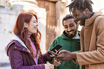 Diverse friends sharing smartphone content, multicultural group laughing outdoors, social media...