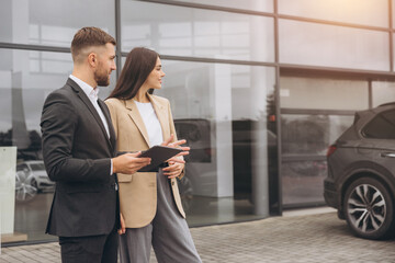 Bearded business man car salesman in grey suit giving explanations on tablet to pretty young woman...
