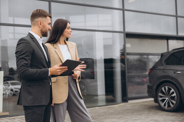 Bearded business man car salesman in grey suit giving explanations on tablet to pretty young woman in beige jacket outside car dealership