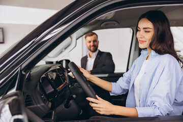Satisfied happy caucasian female client customer woman sitting at the wheel of new car doing test-drive before buying auto while male shop assistant helping her choose it