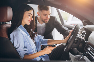 Friendly male manager showing young female customer cars. Beautiful smiling woman sitting inside vehicle in car. Bearded sales consultant working in car dealership.