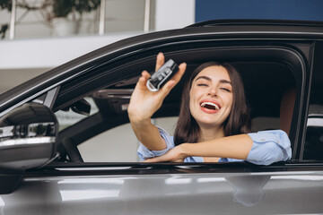 Happy smiling woman sitting in new car and holding keys in car dealership showroom