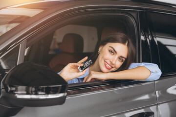 Happy smiling woman sitting in new car and holding keys in car dealership showroom