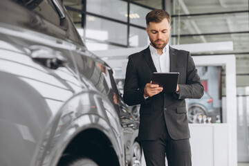 Friendly car seller in grey suit standing in car salon and holding tablet. It's always pleasure to...
