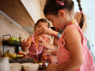 Two children in pink outfits are helping an adult break eggs into a bowl in a modern kitchen, highlighting engagement and bonding. Several ingredients are nearby.