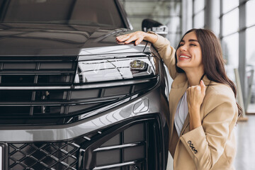 Charming young Caucasian woman hugging hood of new car and show fist and say yes in modern bright...