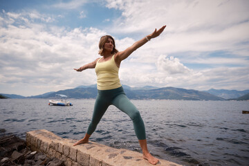 A woman confidently executes a warrior pose in yellow top and green leggings near the sea, with a distant boat and mountains, conveying strength and stability.