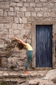 A woman performs a standing back-bend yoga pose by a stone wall, highlighting her flexibility, strength, and the beauty of blending physical activity with ancient structures.