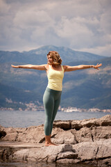 A woman in a yellow top and green leggings stands with arms outstretched on a rocky shore, gazing at the mountains and sea, symbolizing freedom and openness.