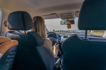 widok z tylnego siedzenia samochodu na kobietę z długimi, jasnymi włosami, która prowadzi pojazd. Kobieta trzyma kierownicę, a w tle widać drogę i jasne niebo przez przednią szybę.  © Clear Emotion