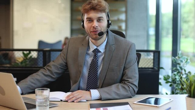 Smiling male call-center operator with headphones sitting at modern office, consulting online information, assistance to the client, looking at camera.