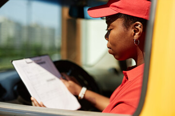 African American woman in red uniform reviewing checklist while sitting in delivery van with focus...