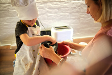 A young aspiring chef, adorned in a white apron and hat, blends with an adult on the side,...