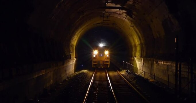 Freight train enters dark tunnel at night in rural area, illuminating tracks with bright headlights during evening journey
