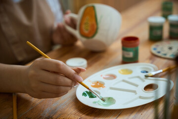 Female hand dipping paintbrush into palette while painting on clay pottery mug