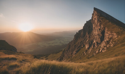 Fototapeta premium A rocky mountain peak juts out against a bright sunset sky, as seen from a grassy hillside below