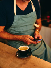 A barista wearing an apron uses a smartphone while a cup of freshly brewed coffee sits on the counter in a modern coffee shop, showcasing a moment of multitasking.