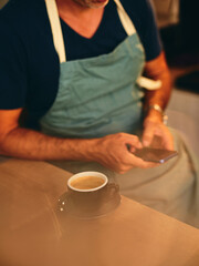 A barista in a blue apron is seen using a smartphone while enjoying a cup of coffee, suggesting a modern, relaxed, and professional coffee shop environment.