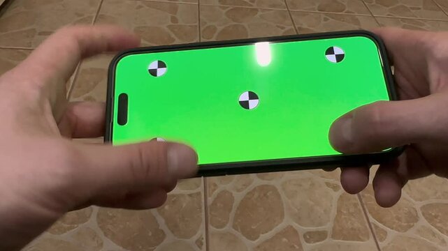 first person view of the hands of a person holding a phone with a green screen with markers and tilting and tapping it periodically