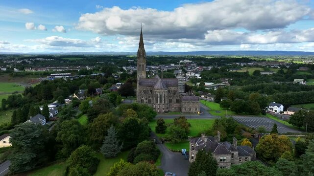 St. Macartan's Cathedral, County Monaghan, Ireland, September 2022. Drone orbits clockwise pushing closer to the Gothic Revival architecture surrounded by green trees and gardens under a overcast sky.
