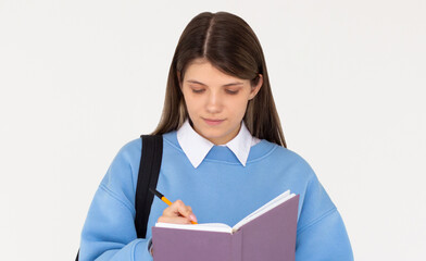 Obraz premium Happy teen student girl wear blue sweatshirt and backpack hold book write down lesson in exercise book isolated on white background Education in university college concept