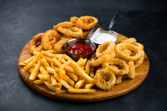 Set of fried onion rings, breaded squid rings, French fries and sauces on a wooden board.
