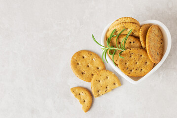 Oval crispy cookies or crackers with rosemary in heart-form bowl  on light textured background, top view with copy space