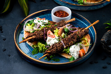 Kebab, barbecued beef on a stick with tomatoes, lettuce, onion, arugula, sour cream, tortilla and red sauce on a plate.