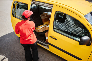 Worker in red uniform loading packages into a yellow delivery van. Scene taken from overhead angle showing interior details of the van and packages being organized