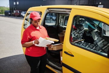 Delivery worker wearing red uniform checking packages beside yellow van in outdoor setting, engrossed in verifying delivery list while holding a package