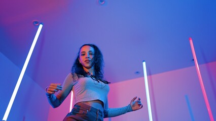 Low angle camera of hispanic dancer looking at camera and dancing to city pop music. Professional attractive hipster moving to hip hop song with purple led or neon light. Stylish cloth. Regalement.