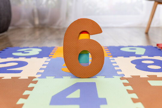 r&oacute;jwymiarowa cyfra "6" stoi pionowo na kolorowej macie piankowej z puzzli. W otworze cyfry widoczne są fragmenty kolorowych klock&oacute;w lub element&oacute;w maty, co sugeruje, że jest to wyjmowany element. 