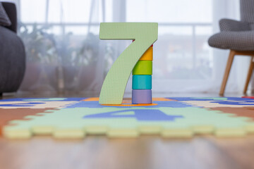 cyfra 7 stoi pionowo na kolorowej macie piankowej z puzzli dla dzieci. Liczba stoi na macie piankowej a za nią stoją klocki w różnych kolorach, koncept edukacji dziecka.  © Clear Emotion