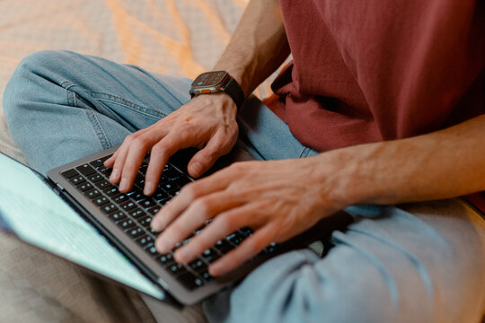 A man is typing on a laptop while sitting on a bed at home. Close-up of his hands, an unrecognizable person