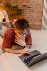 A man in casual clothes with earphones uses a laptop lying on a bed at home. He is talking on video...