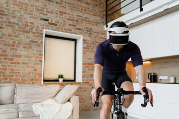 A man in sportswear and a VR headset trains on a bicycle mounted on a stand at home. Daylight