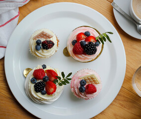 Cupcakes and mini pavlova cakes decorated with fresh berries and white chocolate on a large white plate, a cup of coffee and a cloth napkin on wooden table. Summer fruit cakes flat lay.
