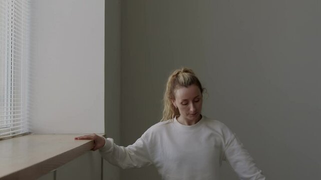 Young talented ballerina using window sill as barre while practicing classical ballet poses with feet in first position in studio