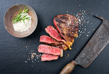 Traditional barbecue dry aged rib-eye beef steak with salt in a bowl sliced as top view on a black...