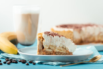 Slice of banoffe pie filled with banana and dulce de leche and sprinkled with grated chocolate. Banana cake on white plate with glass of coffee on turquoise summer background