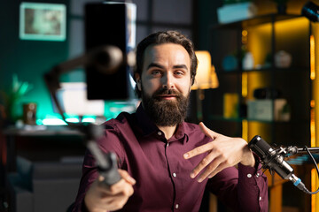 Content creator talking in front on cellphone attached to selfie stick in home studio interior,...