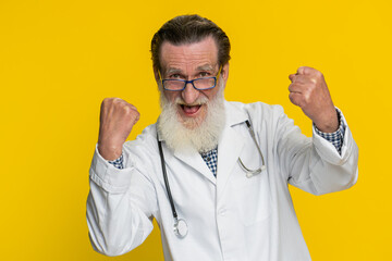 Elderly doctor cardiologist old man celebrating success victory, winning, achievement, good positive news, triumph, succeed. Apothecary pharmacy grandfather champion isolated on yellow background