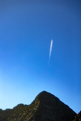  Airplane flying high in the clear blue sky above a rugged mountain peak, leaving a white contrail...