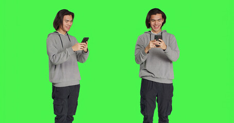 Guy texting messages on mobile phone standing against greenscreen backdrop, using social media app to chat remotely with friends. Asian joyful adult messaging on smartphone.