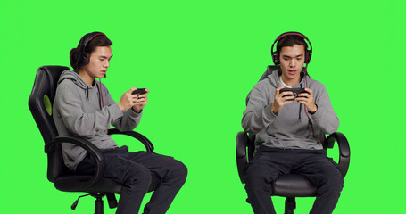 Asian young man plays mobile games over greenscreen, holding smartphone to play online multiplayer competition. Gamer using phone device to have fun with new rpg gameplay on chair.