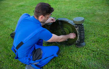 Man in blue overalls is carefully adjusting a lawn mower blade and cleaning while kneeling on a well-kept lawn. The importance of proper equipment maintenance for effective lawn care.