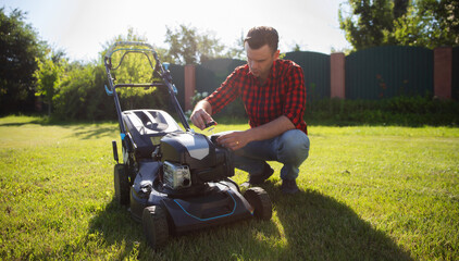 Fototapeta premium A man in a red plaid shirt kneels on a lawn, performing maintenance on a lawn mower under the bright sunlight. Hands-on outdoor work and the importance of tool care for effective lawn maintenance.