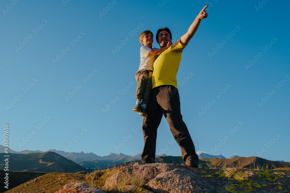 Wall mural Father and son standing on huge stone in the mountains at sunset light and pointing hand