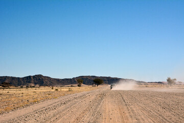 A car drives towards the hills on a dirt road in a desert area in perspective under a clear blue sky